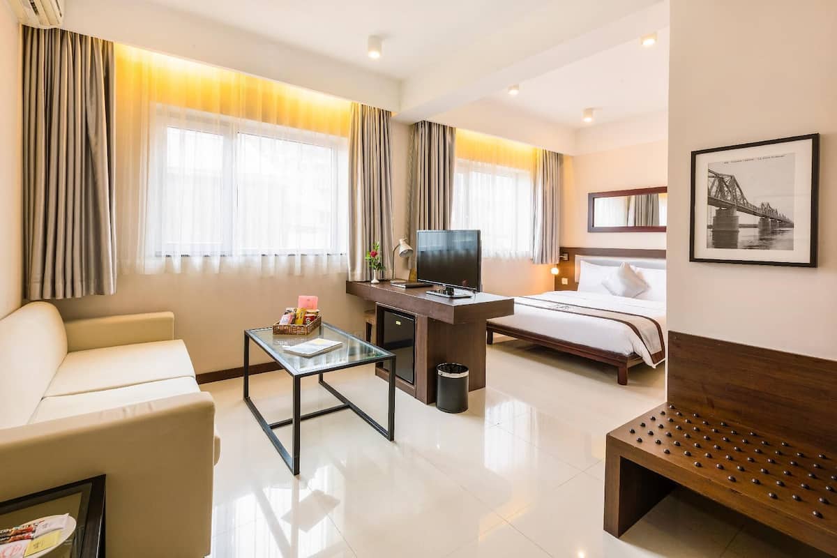 A hotel room with a bed, a desk with a TV, a sofa, a coffee table, and a bench.
