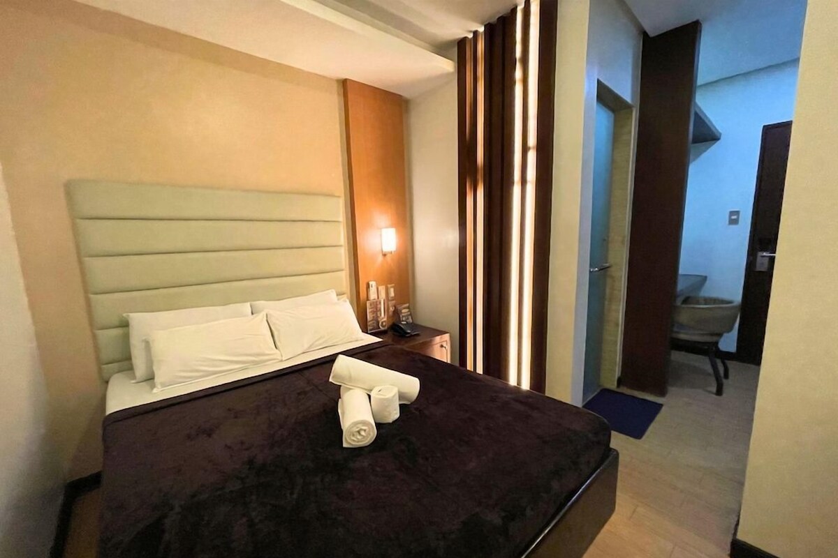 A hotel room with a bed, a nightstand, a lamp, and a view of a hallway.