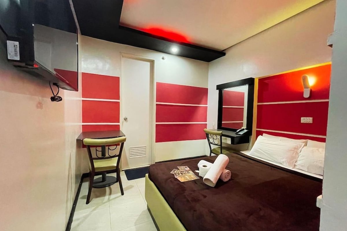 A hotel room with a bed, a desk, a chair, a mirror, and a red accent wall.