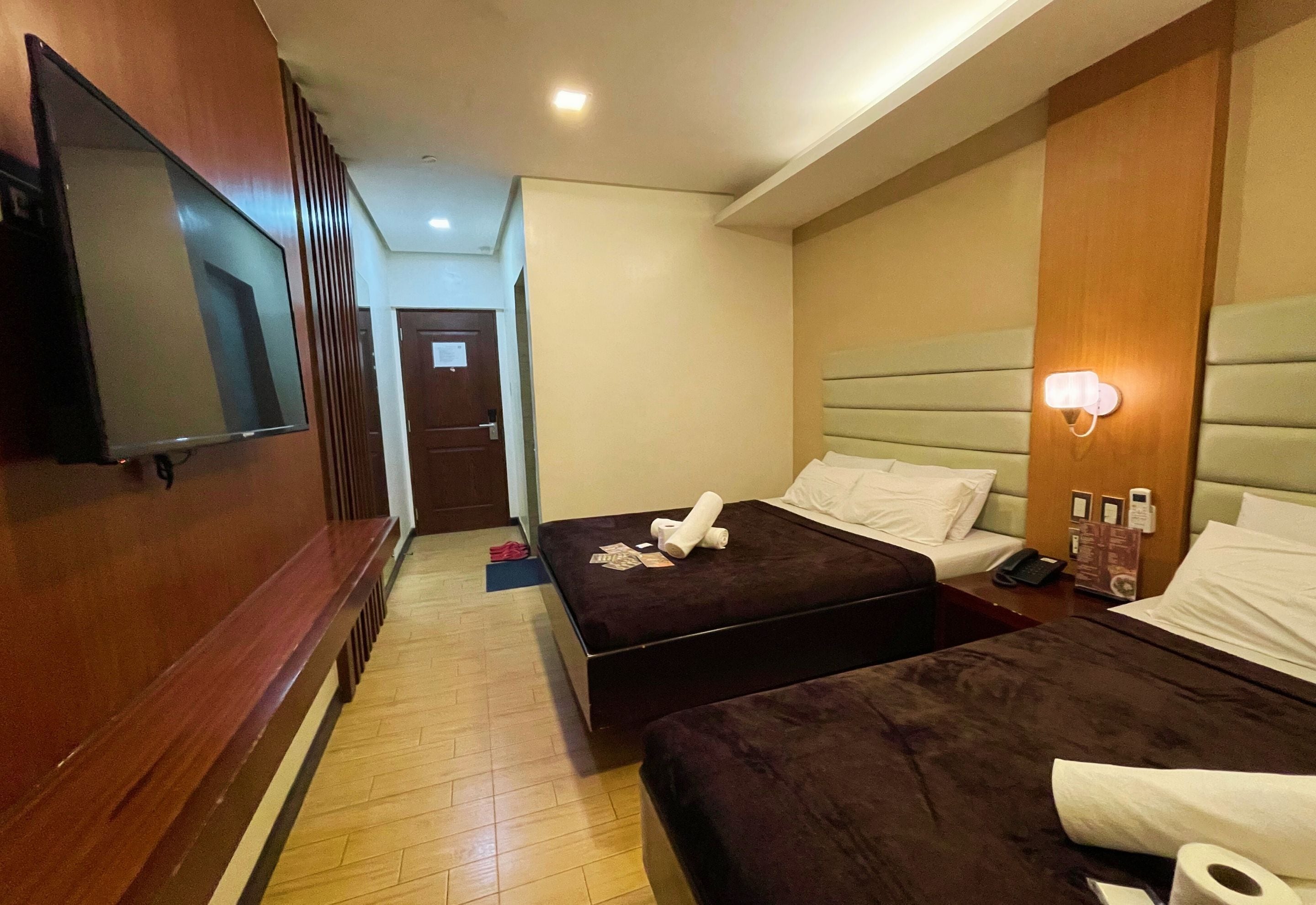 A hotel room with two beds, a TV, a bedside table with a phone and a lamp, and a wooden panel wall.