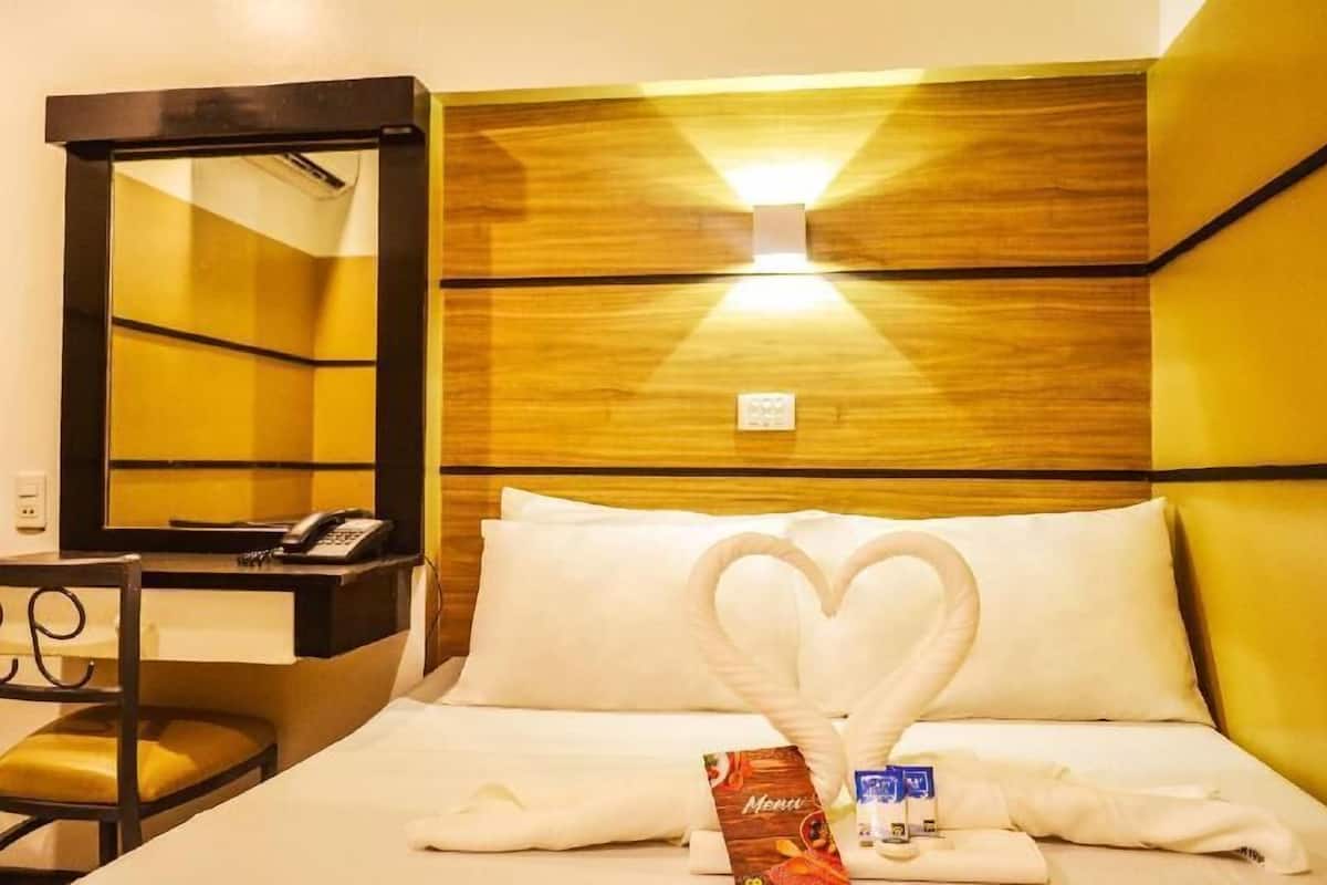 A hotel room with a bed, a desk, a chair, a mirror, and a heart-shaped towel arrangement.