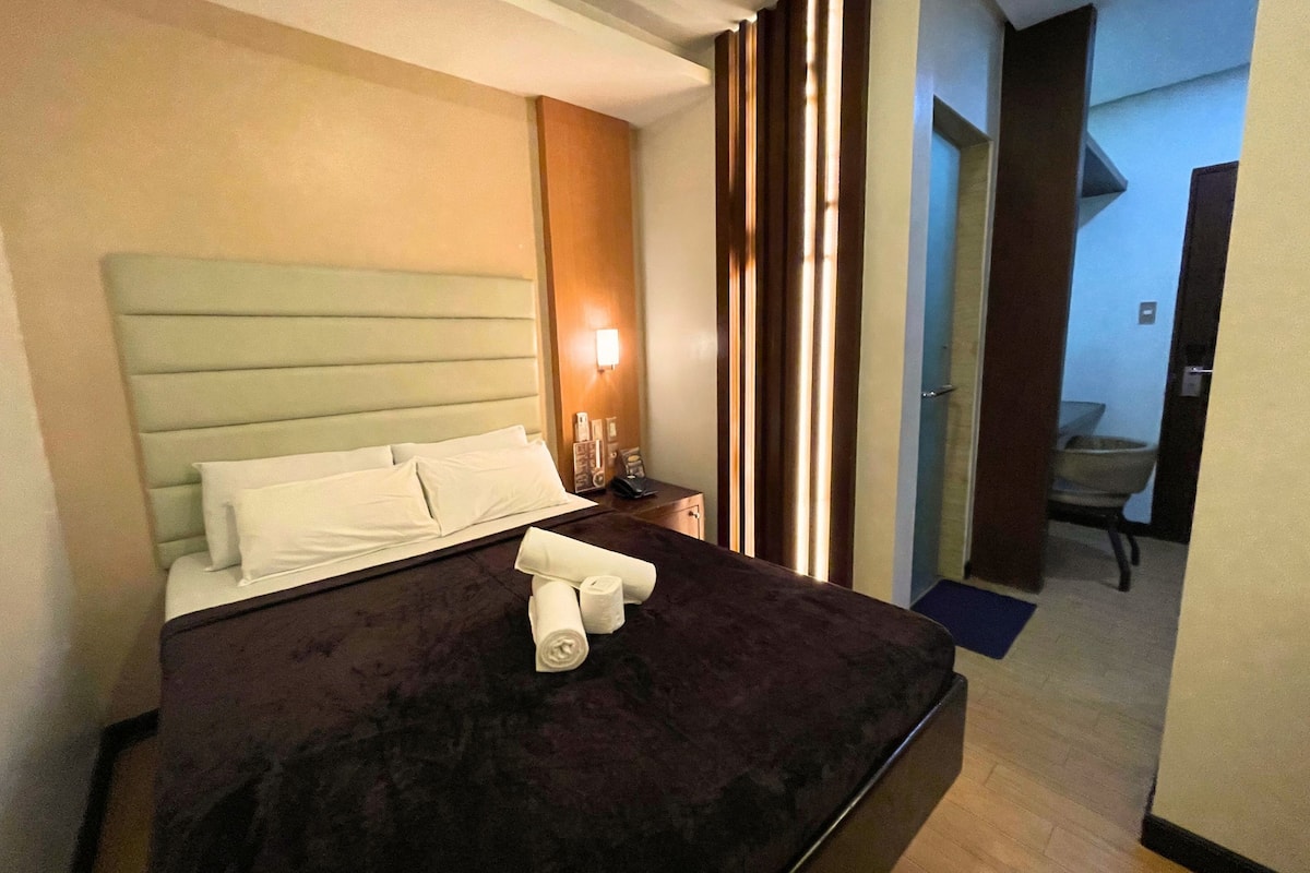 A hotel room with a bed, a nightstand, a lamp, and a view of a bathroom.