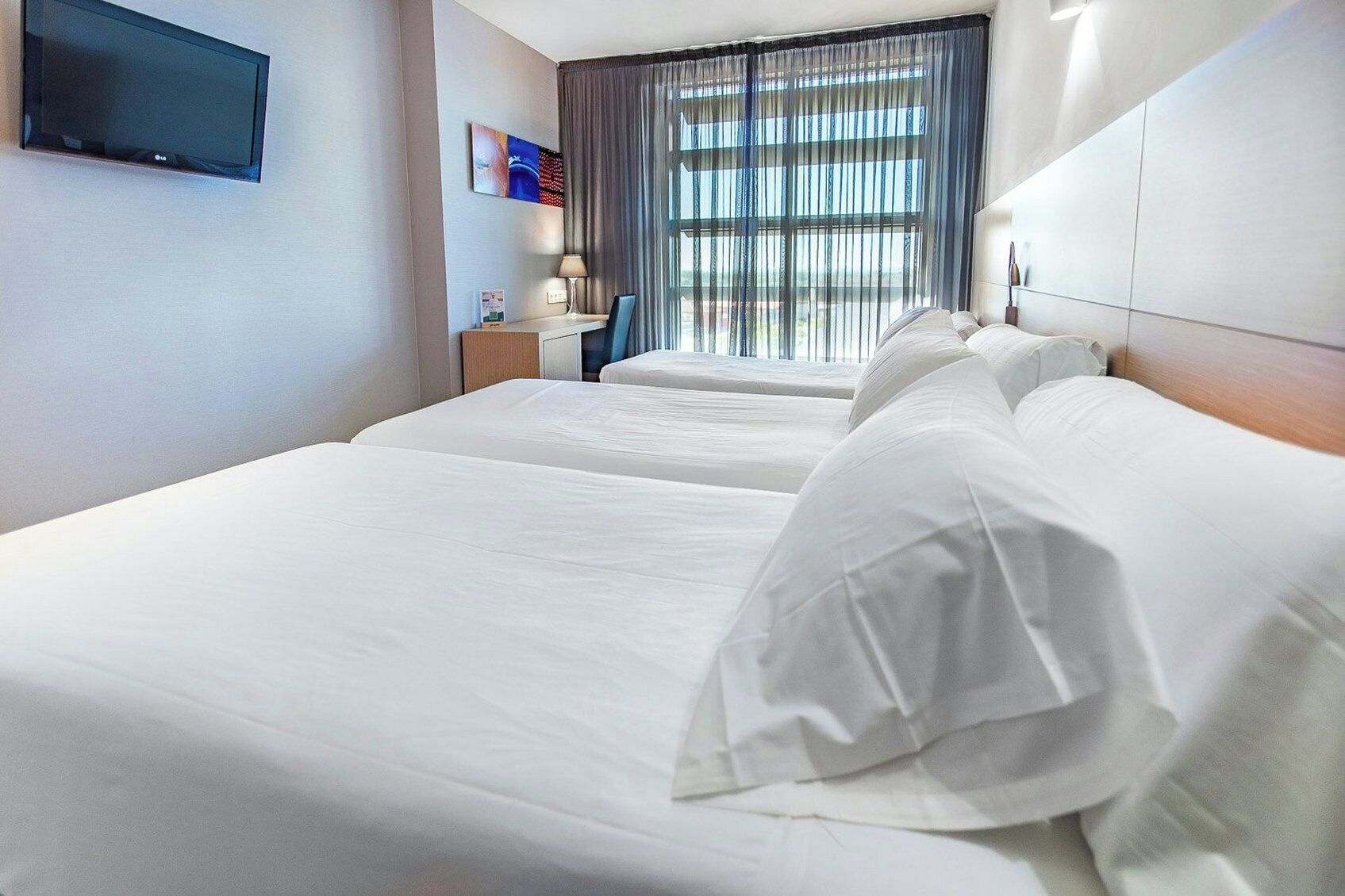 A hotel room with two beds, a flat-screen TV mounted on the wall, a desk with a lamp, and a window with curtains.