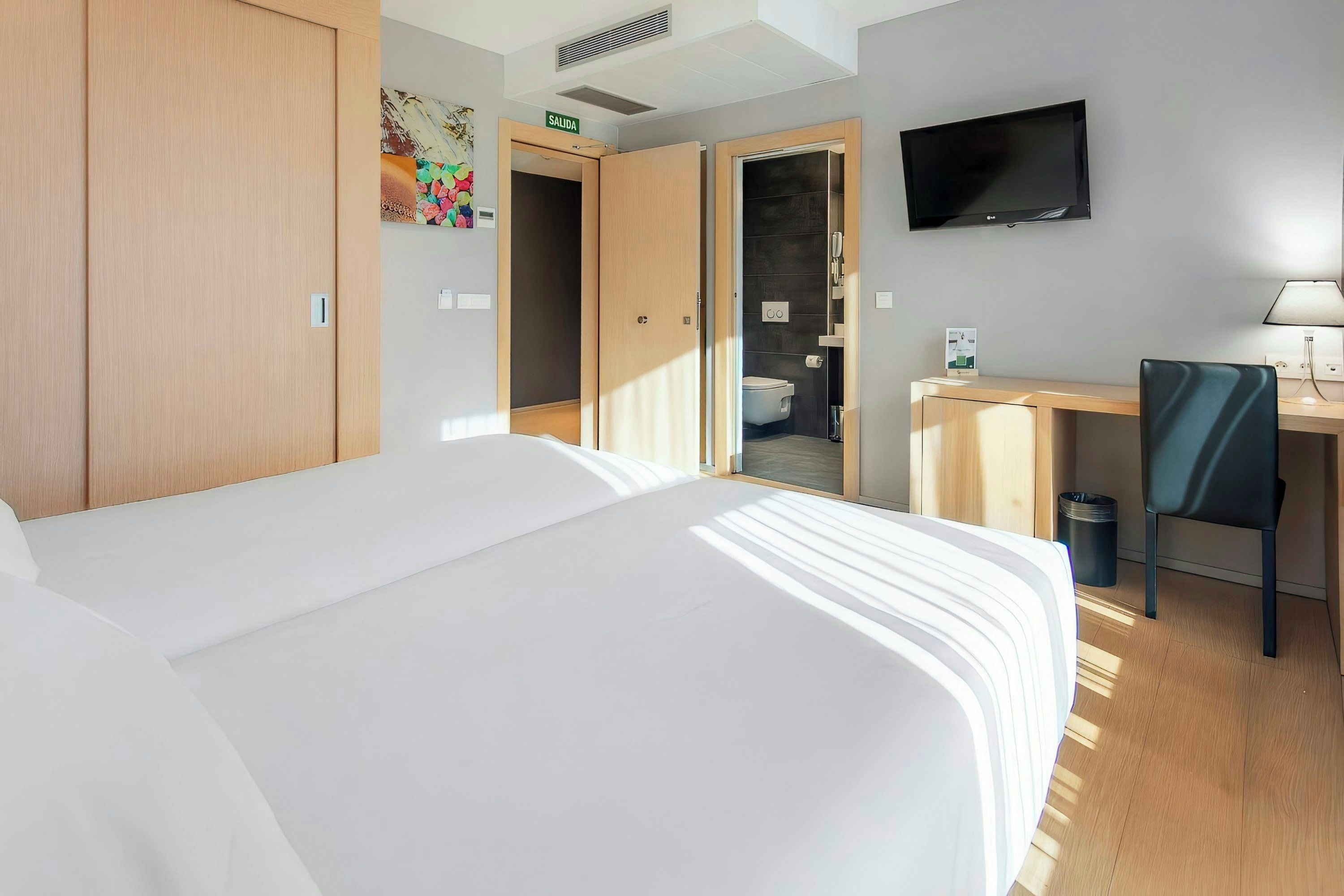 A hotel room with a bed, a desk, a chair, a TV, and a bathroom visible through an open door.
