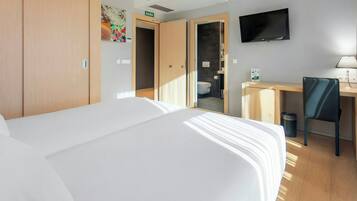 A hotel room with a bed, a desk, a chair, a TV, and a bathroom visible through an open door.