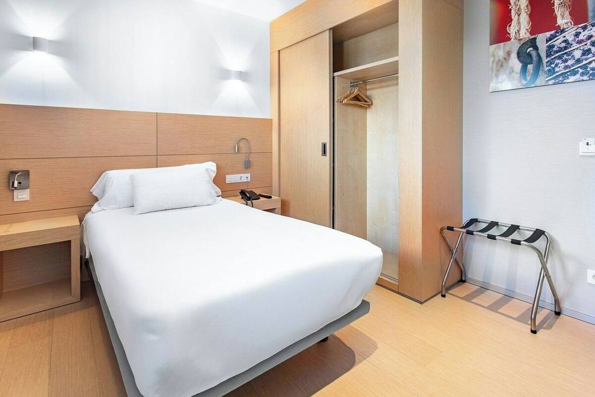 A hotel room with a bed, bedside table, wall-mounted light, and a coat rack.
