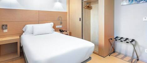 A hotel room with a bed, bedside table, wall-mounted light, and a coat rack.