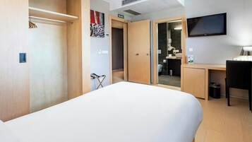 A hotel room with a bed, a desk, a chair, a TV, and a bathroom visible through an open door.