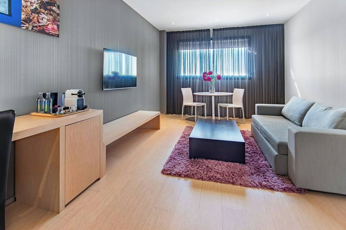 A modern living room with a sofa, a coffee table, a TV, a dining area with chairs, and a desk with a computer and a coffee machine.