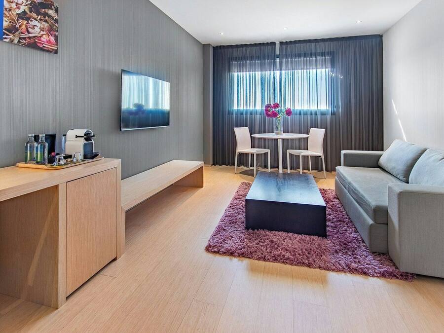 Uma sala de estar moderna com sofá, mesa de centro, televisão, área de jantar com cadeiras, e escrivaninha com computador e máquina de café.