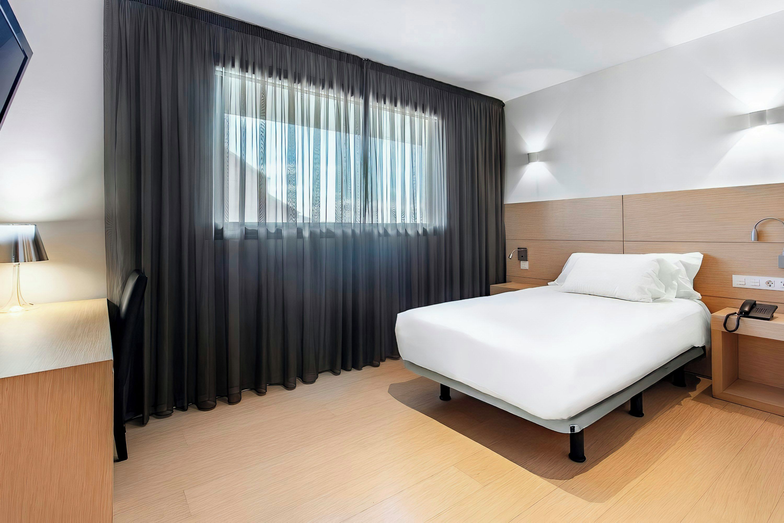 A hotel room with a bed, bedside table, lamp, and a window with curtains.