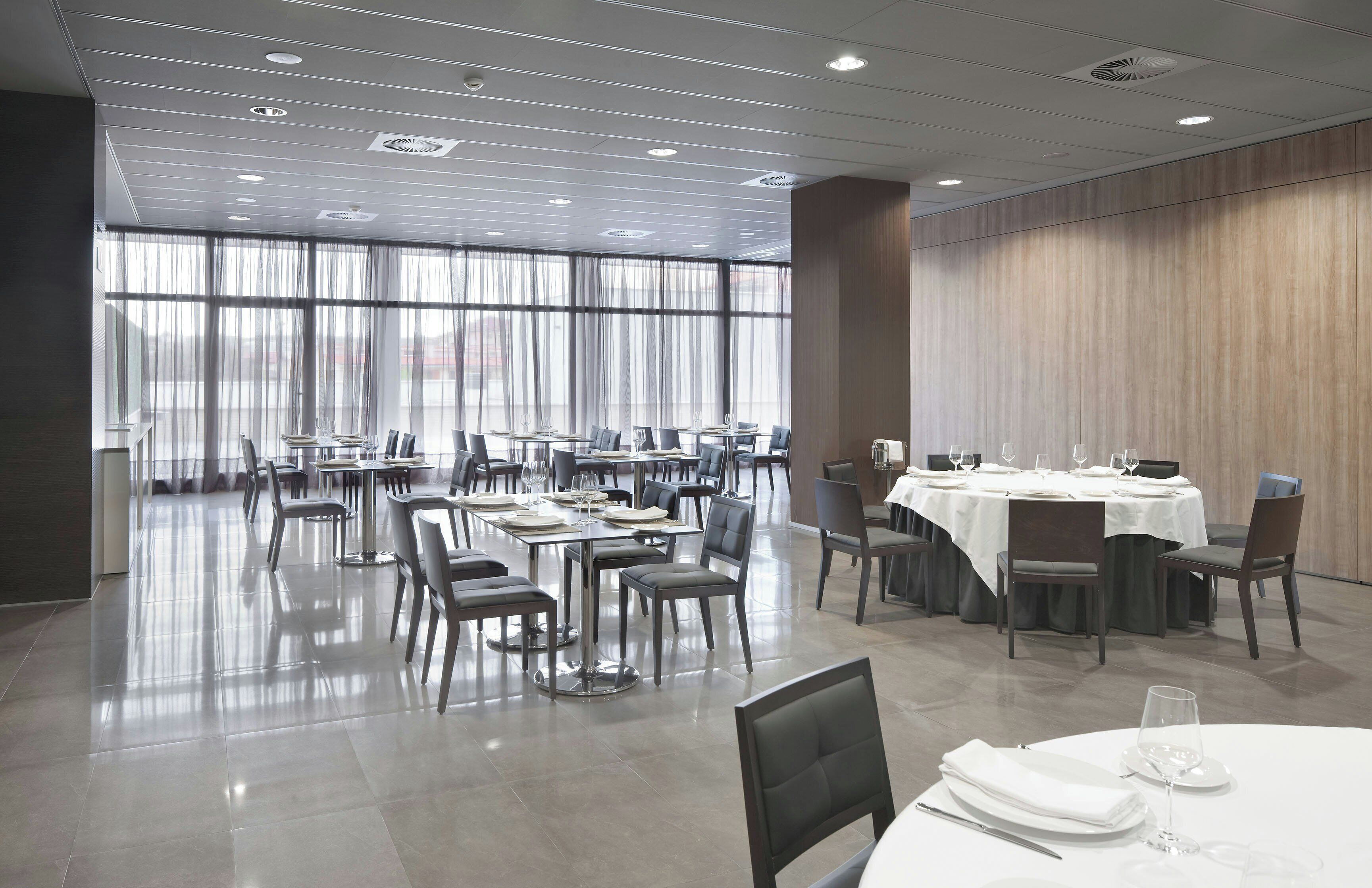A modern dining area with tables set for meals, large windows with curtains, and a polished floor.