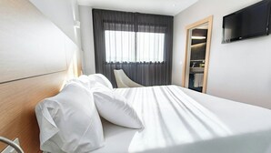 Una cama bien hecha con sábanas blancas, una mesita de noche, un televisor montado en la pared y una ventana con cortinas.