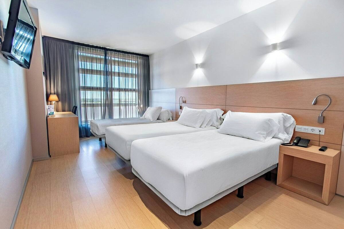 A hotel room with two beds, a desk, a TV, and a window with curtains.