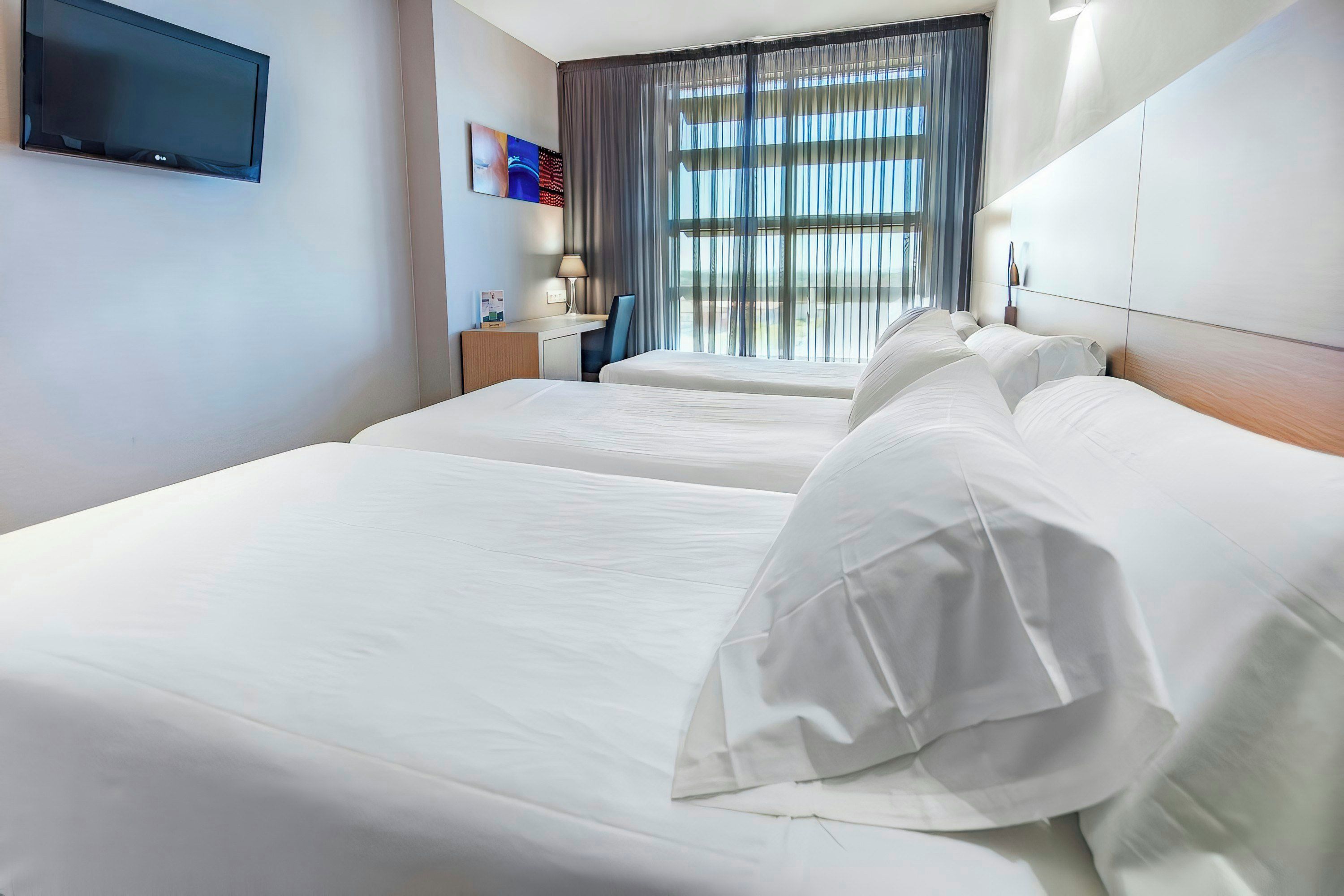 Habitación de hotel con dos camas, un televisor de pantalla plana montado en la pared, un escritorio con lámpara y una ventana con cortinas.