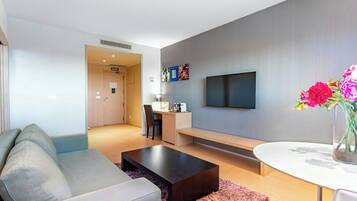 A modern hotel room with a sofa, a coffee table, a TV, a desk, and a chair.