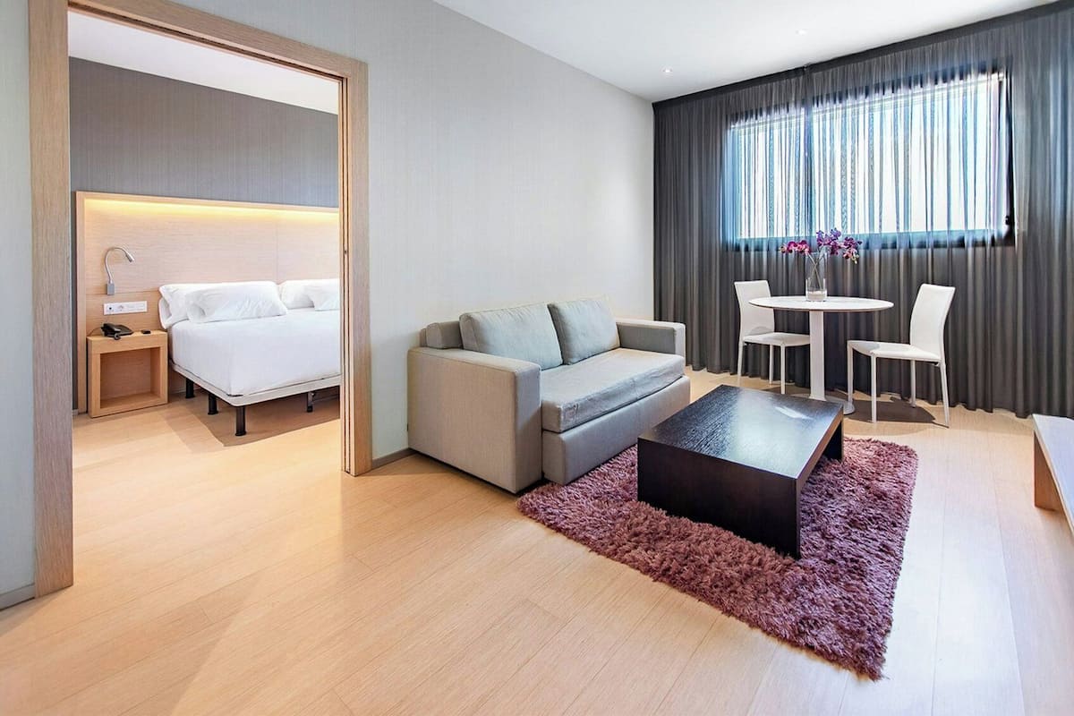 A modern hotel room with a bed, sofa, coffee table, and a dining area with chairs and a table.