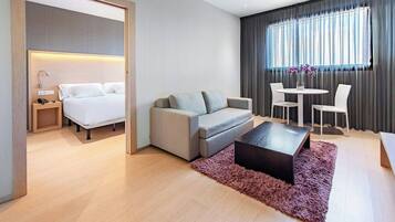 A modern hotel room with a bed, sofa, coffee table, and a dining area with chairs and a table.