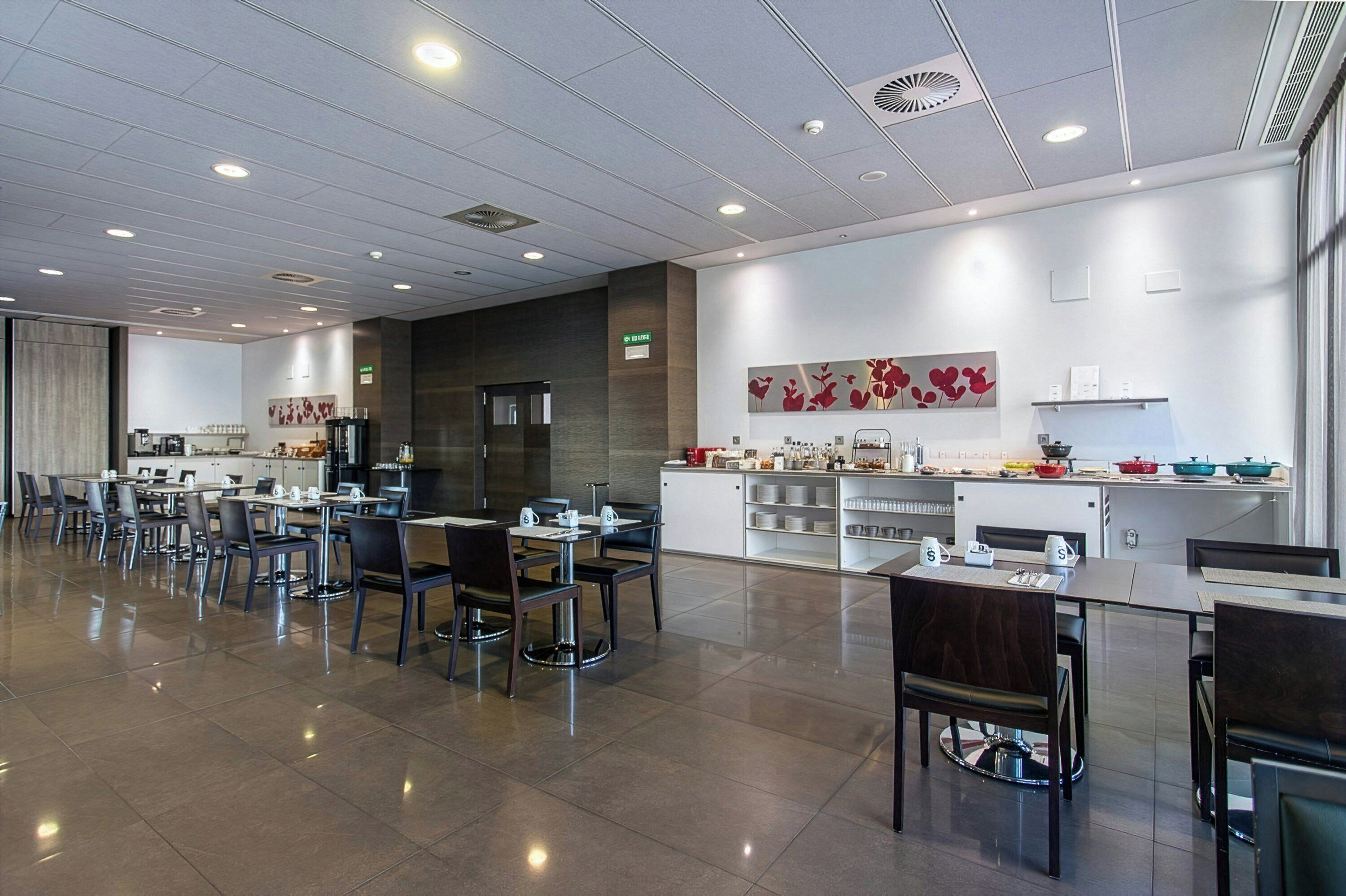 Un comedor moderno con varias mesas y sillas, una zona de cocina con electrodomésticos, y una obra de arte decorativa en la pared.
