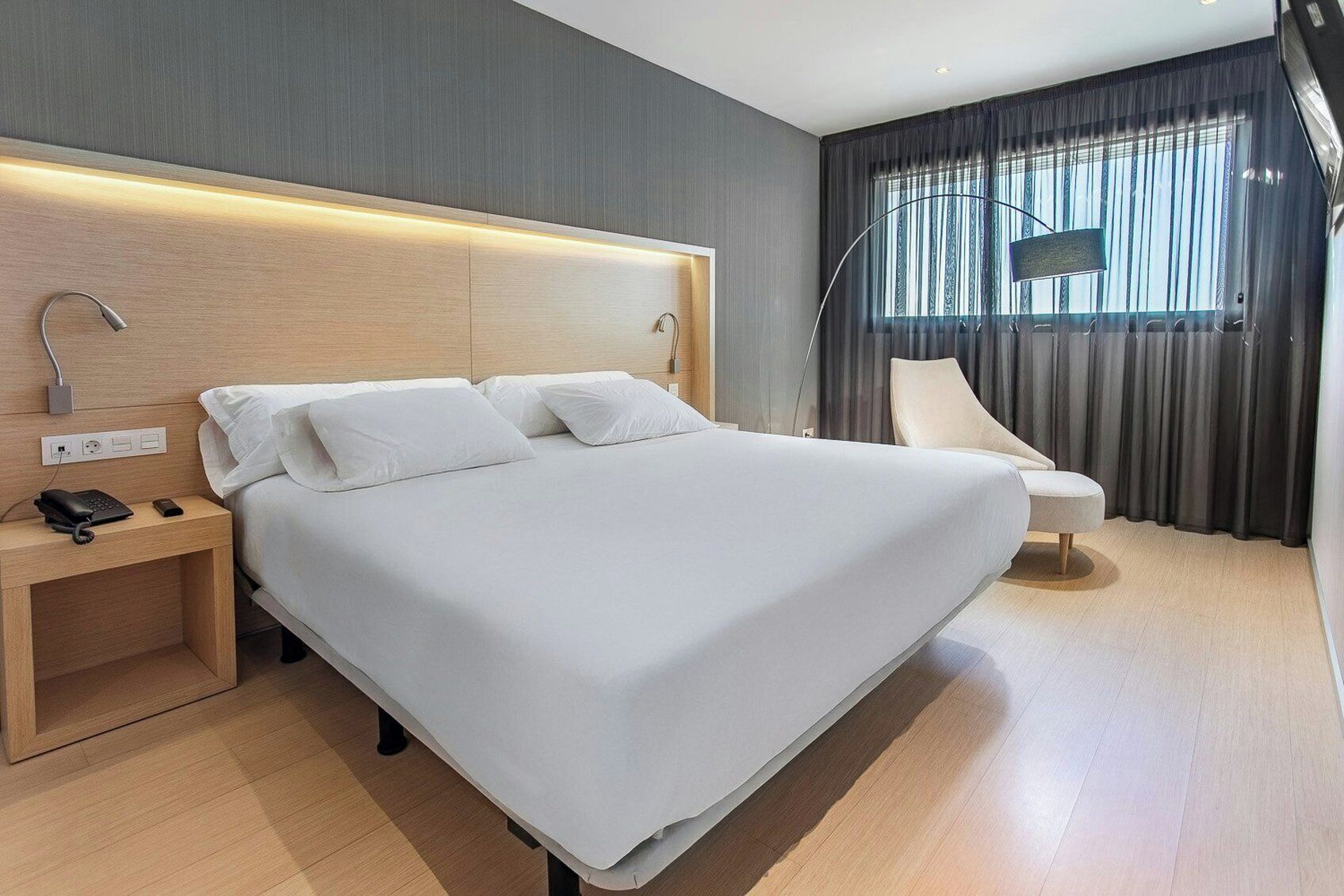 A modern hotel room with a large bed, bedside table with a phone, and a chair.