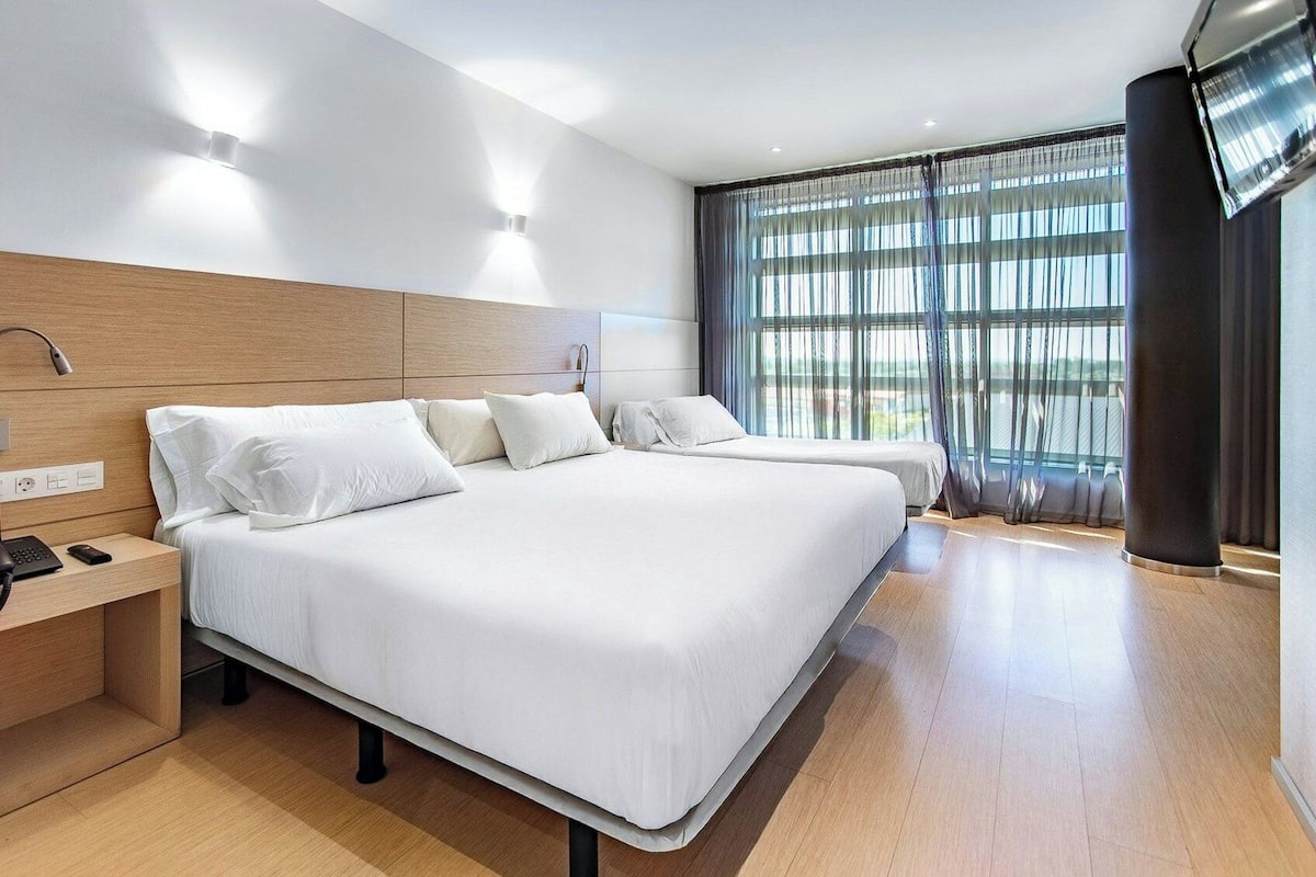 A modern hotel room with a large bed, bedside tables, a flat-screen TV, and a large window with curtains.