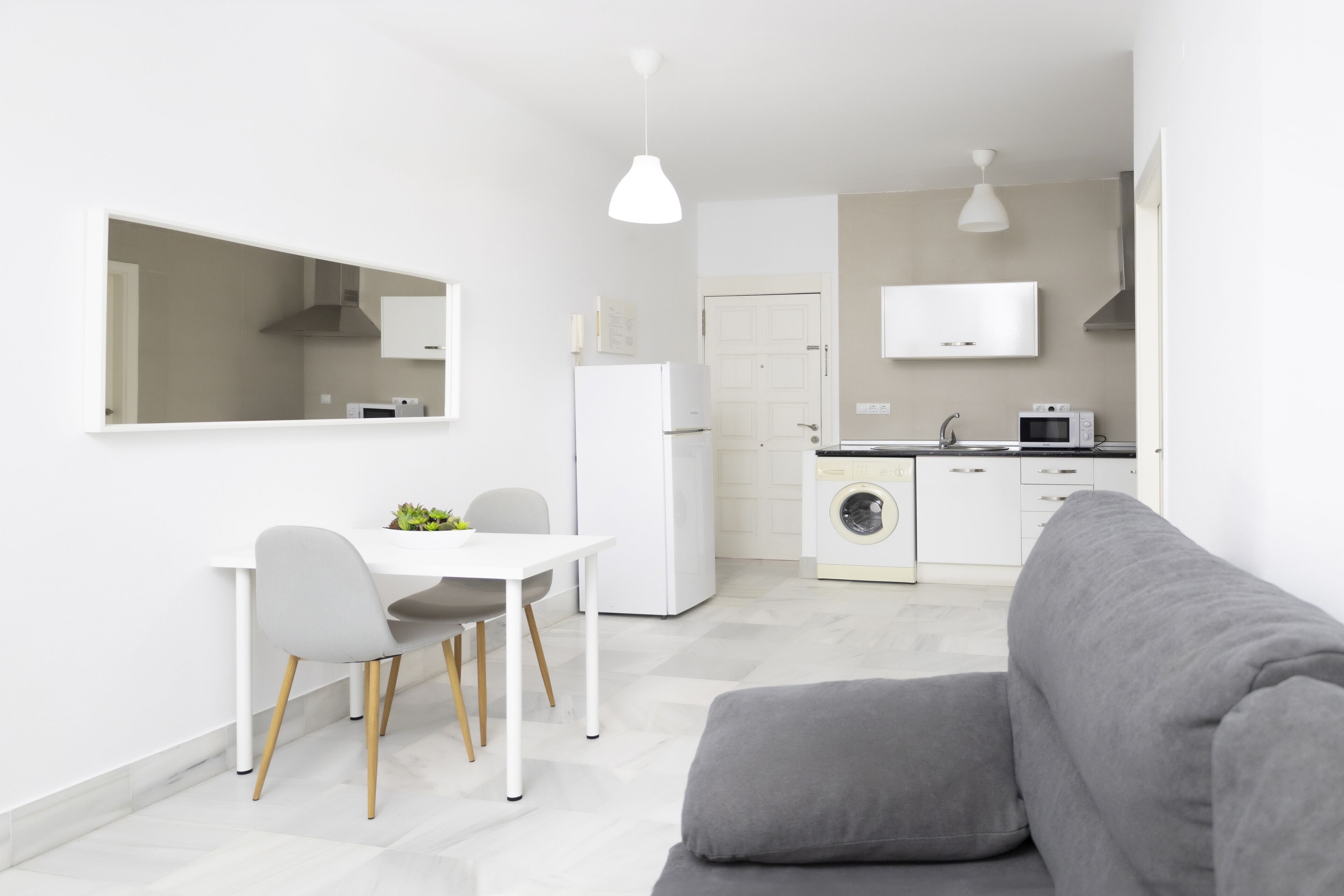 A modern kitchen with white appliances, a dining area with a white table and chairs, and a grey sofa in the foreground.