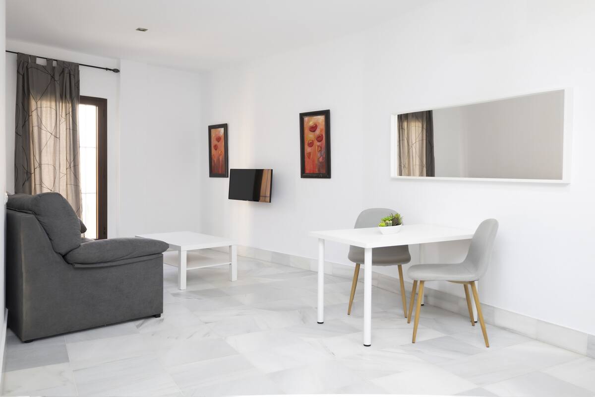 A modern living room with a grey sofa, a white dining table with two chairs, and a large mirror on the wall.
