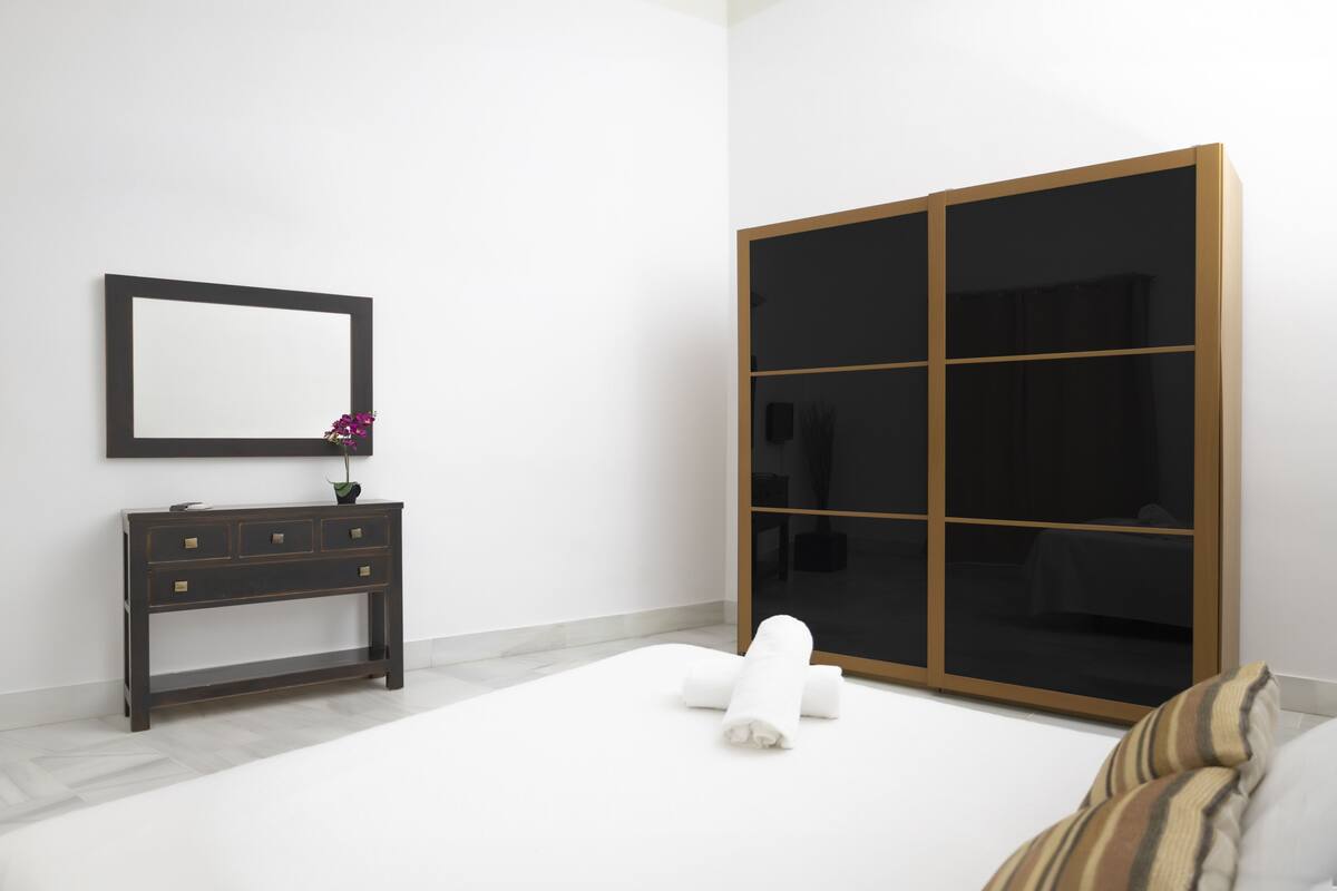 A minimalist bedroom with a large wardrobe, a bedside table with a mirror, and a neatly made bed.