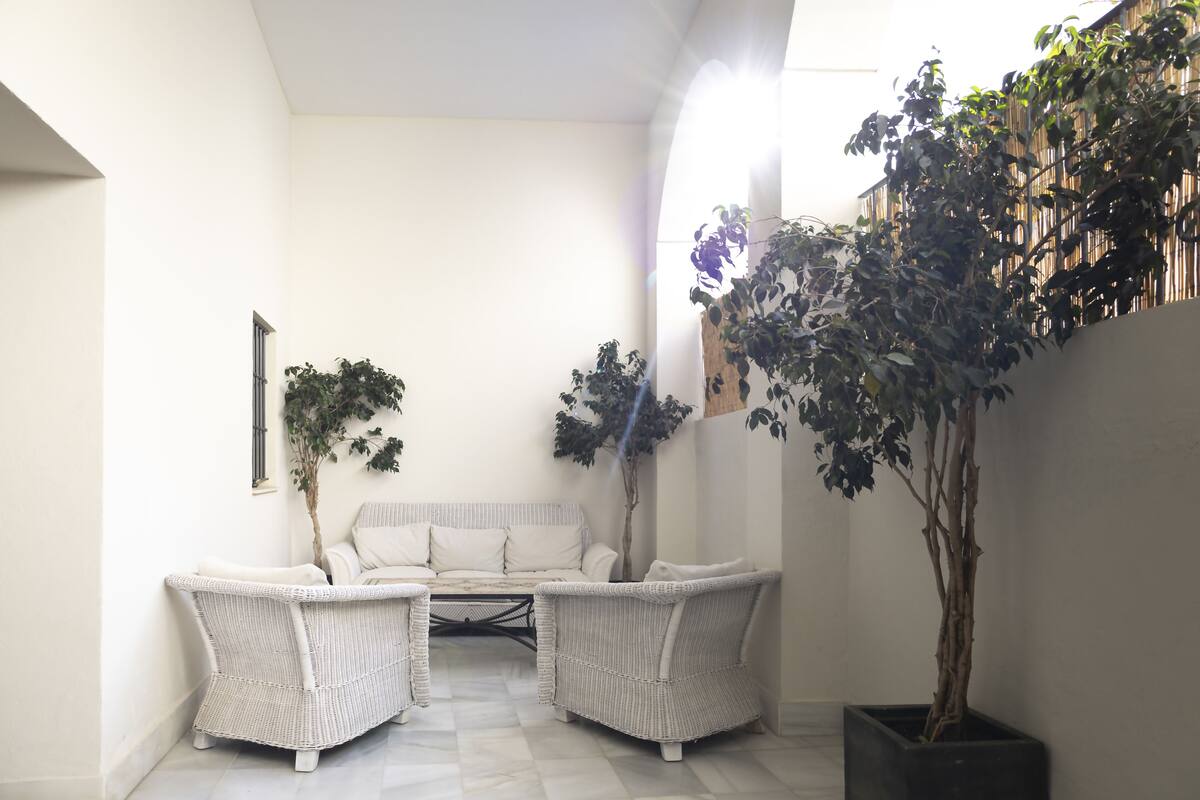 A white wicker sofa set in a corner with a tiled floor, a potted tree, and a wall-mounted shelf.