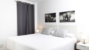 Une chambre à coucher avec un grand lit, deux coussins gris à motifs, une tête de lit blanche, une table de chevet avec une lampe et deux œuvres d’art représentant des éléphants, accrochées au mur.
