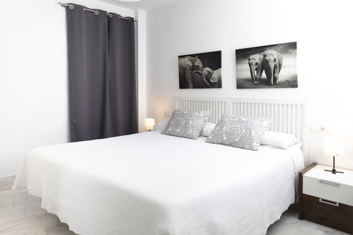A bedroom with a large bed, two gray patterned pillows, a white headboard, a nightstand with a lamp, and two wall-mounted elephant artwork.