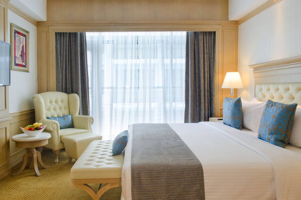 Ein Hotelzimmer mit einem großen Bett, einer Sitzecke mit Sessel und Hocker, einem kleinen Tisch mit Obst und einem an der Wand befestigten Fernseher.