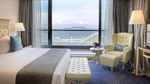 Une chambre d’hôtel dotée d’un grand lit, d’un coin salon avec un fauteuil et un pouf, d’une petite table et offrant une vue sur la ville et l’eau grâce à de grandes fenêtres.
