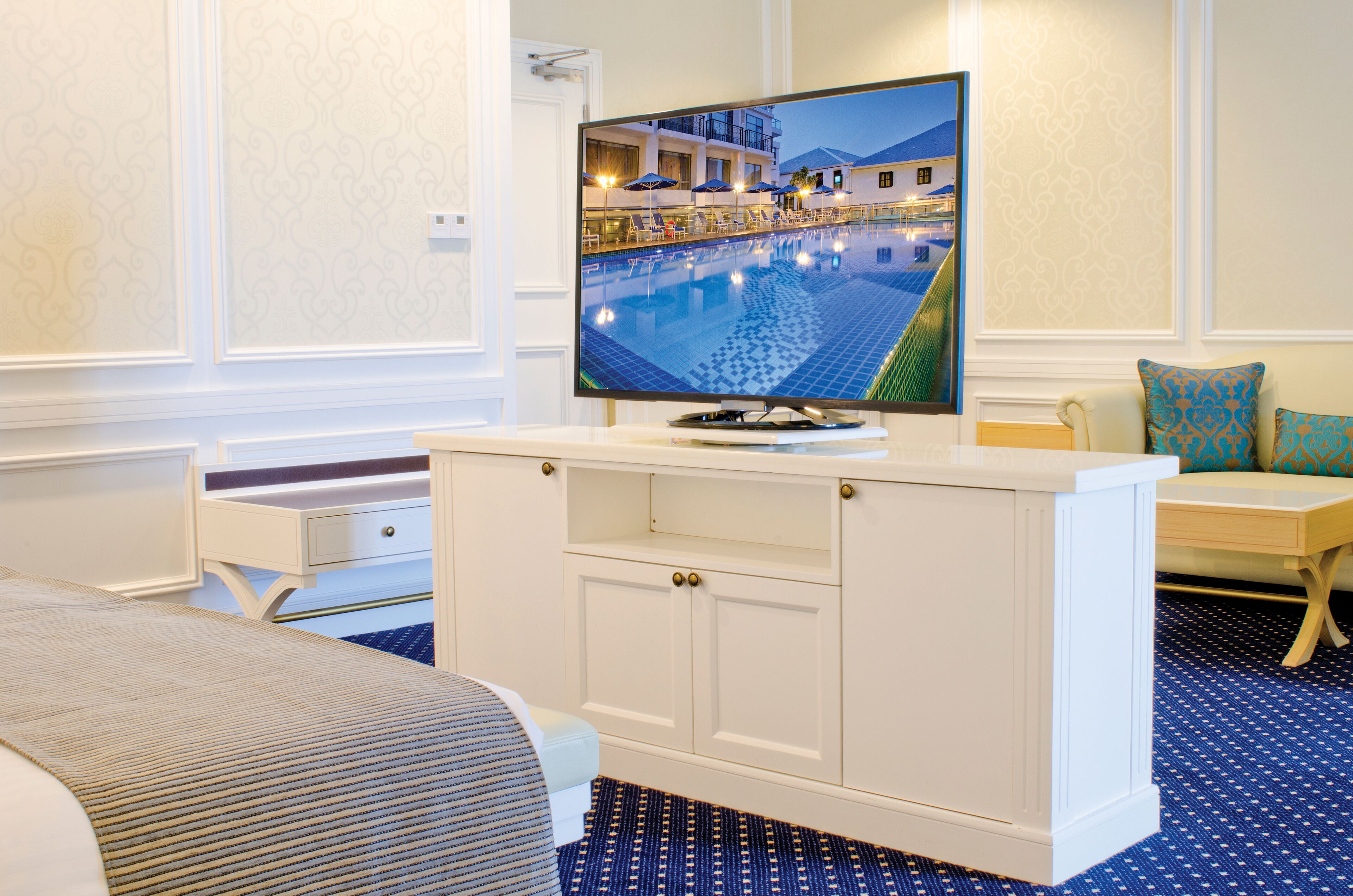 Ein Hotelzimmer mit großem Fernseher, der einen Blick auf einen Pool zeigt, einem weißen Schminktischchen, einem Bett und einem blau gemusterten Teppich.