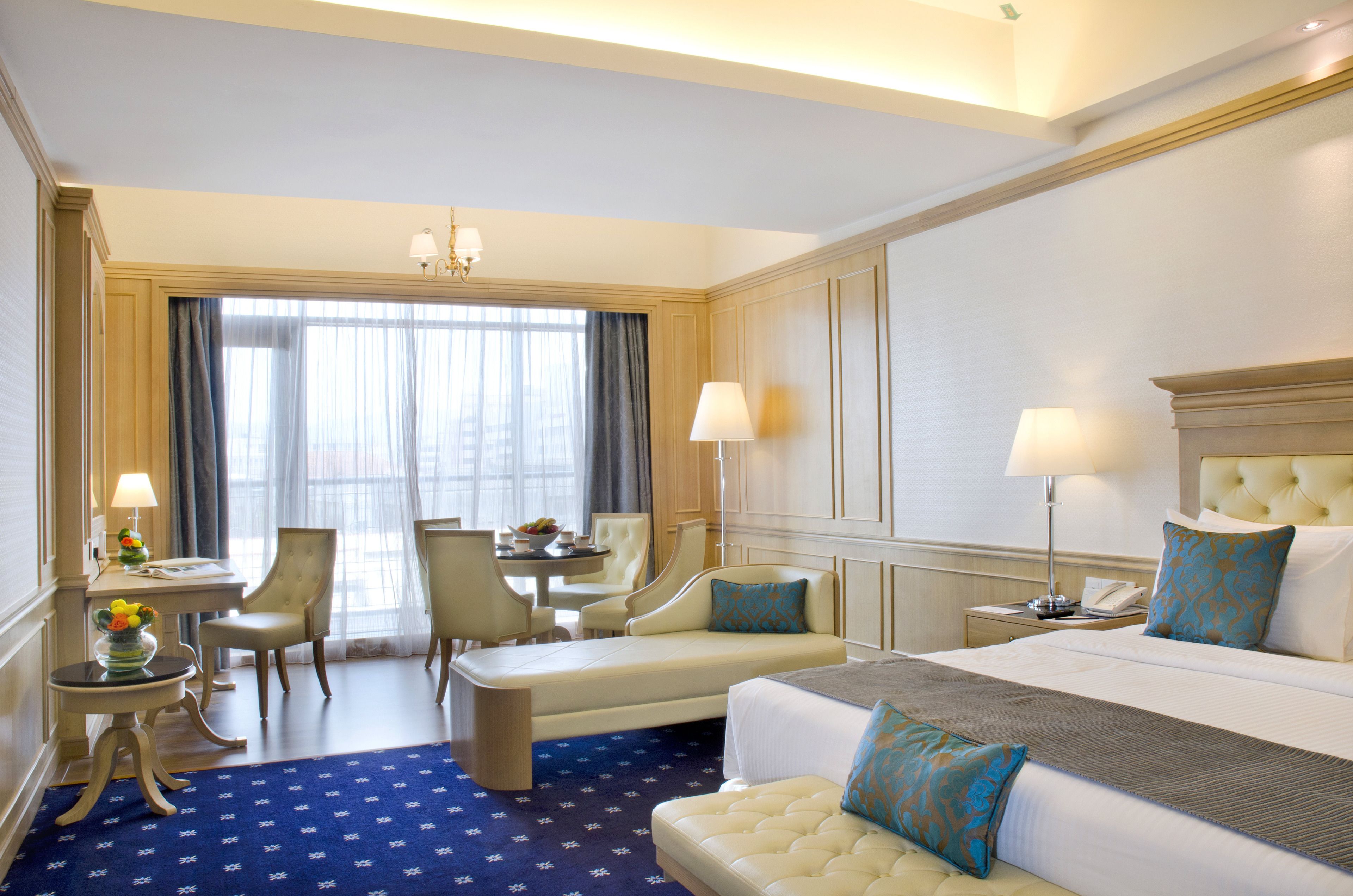 A hotel room with a large bed, a sofa, a dining table with chairs, and a small desk with a lamp.