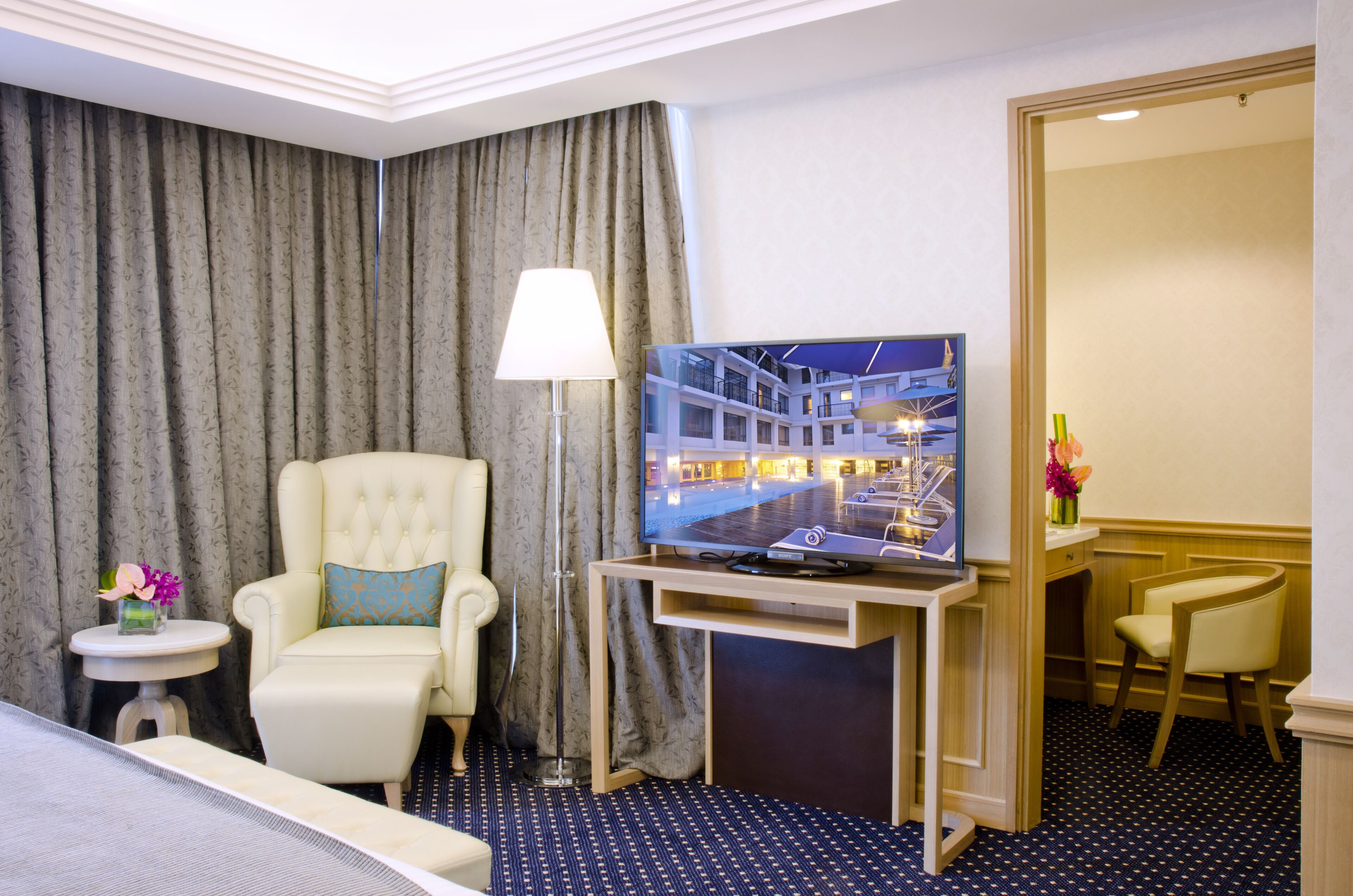 A hotel room with a bed, a TV on a wooden stand, a chair, a desk, and a large window with curtains.