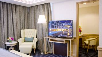 A hotel room with a bed, a TV on a wooden stand, a chair, a desk, and a large window with curtains.