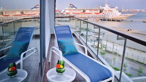 Un balcon avec deux fauteuils et une petite table, donnant sur un port où sont amarrés des navires de croisière et sur le paysage urbain.