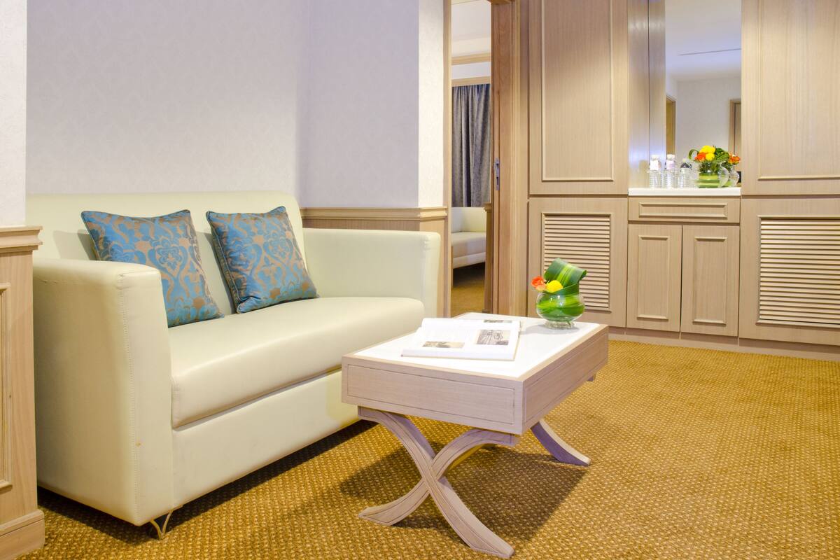 Ein Hotelzimmer mit einem weißen Sofa, einem kleinen weißen Beistelltisch mit Blumen in einer Vase und einem Spiegel über einem Holzschminktisch.