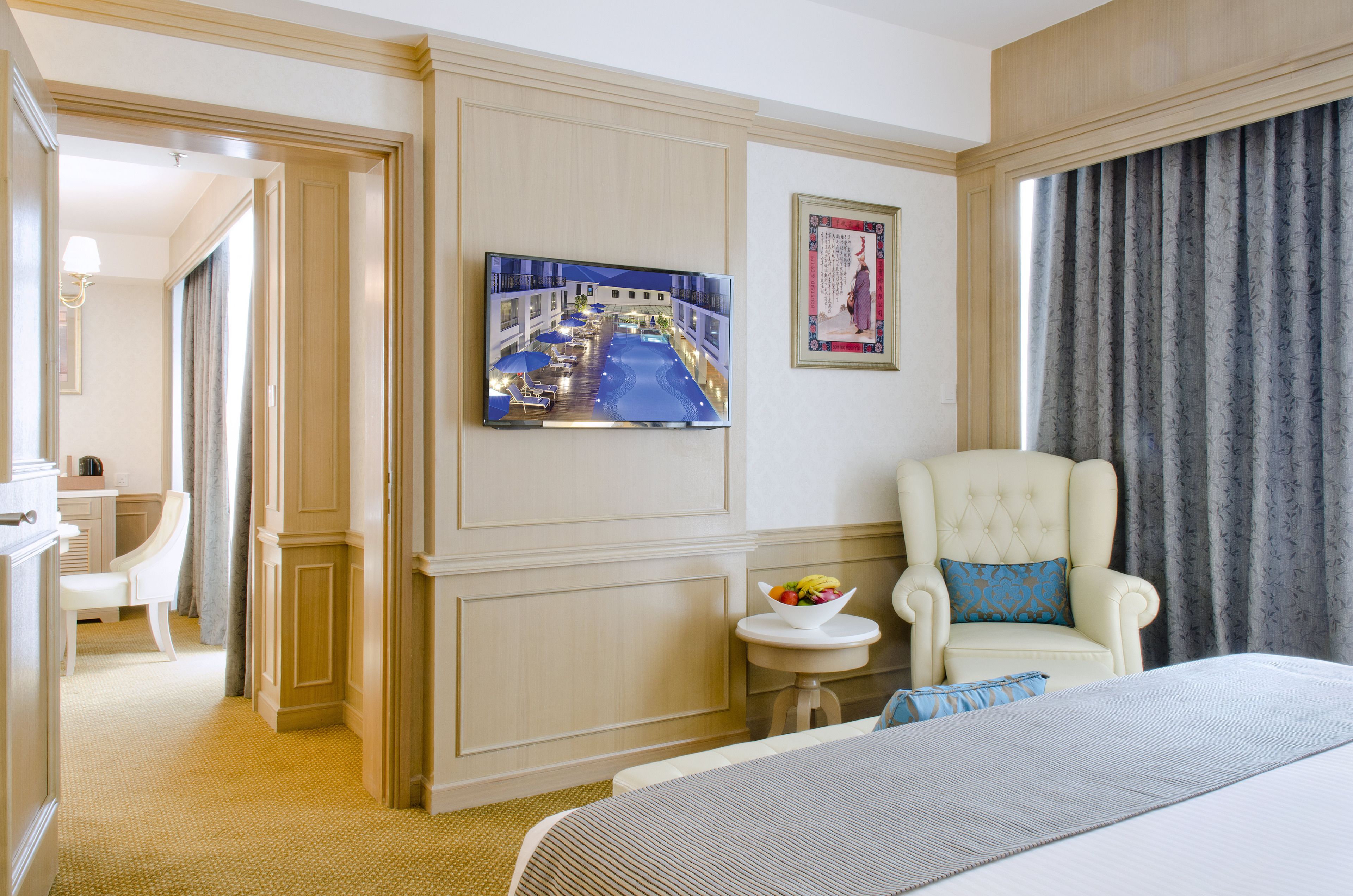 A hotel room with a bed, a television mounted on the wall, a chair with a pillow, a small table with a bowl of fruit, and a framed picture on the wall.
