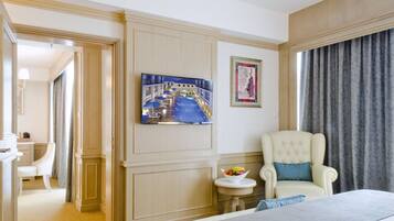 A hotel room with a bed, a television mounted on the wall, a chair with a pillow, a small table with a bowl of fruit, and a framed picture on the wall.