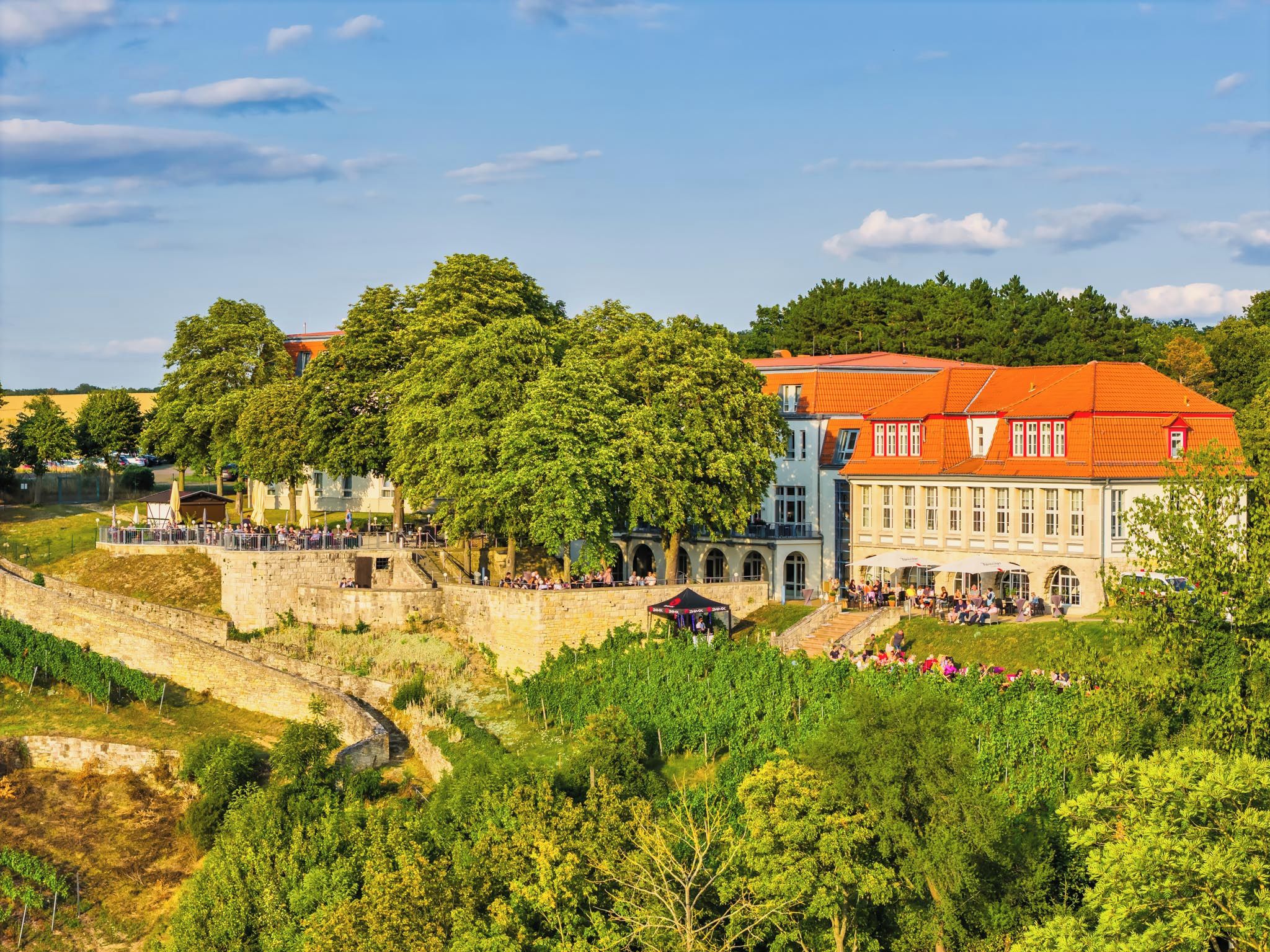 A historic building with a red roof, surrounded by lush greenery and a stone wall.