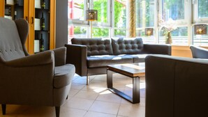 Een moderne woonkamer met een bank, fauteuils en een salontafel. Een boekenkast bevat boeken en decoratieve objecten. Grote ramen zorgen voor natuurlijk licht en uitzicht op de omgeving.
