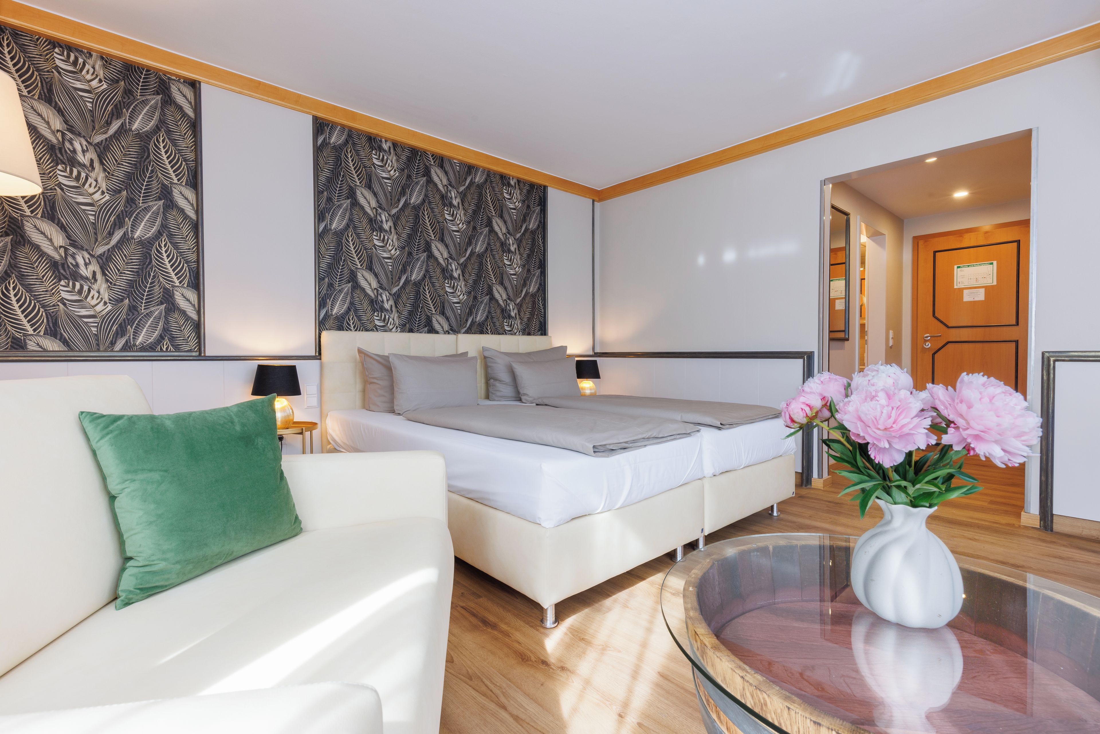 A modern bedroom with a bed, a white sofa, a green pillow, a vase with flowers, and a mirror.