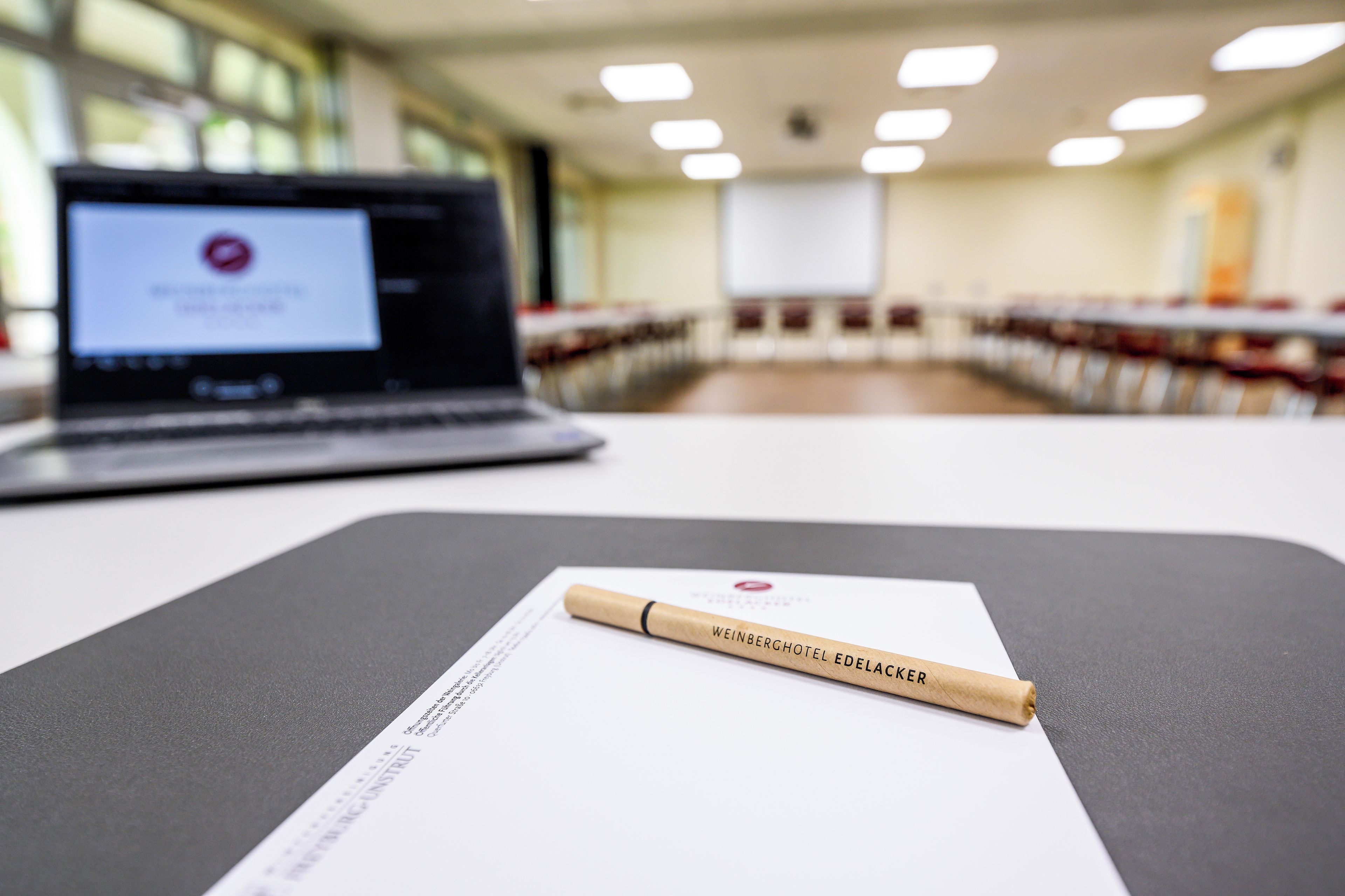 Ein Laptop auf einem Tisch mit einem Dokument und einem Stift, in einem Raum mit Reihen von Stühlen und einer Projektionsleinwand im Hintergrund.