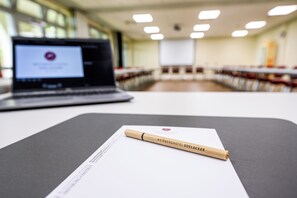 Un ordinateur portable est posé sur une table, accompagné d’un document et d’un stylo, dans une pièce où l’on aperçoit des rangées de chaises et, au fond, un écran de projecteur.