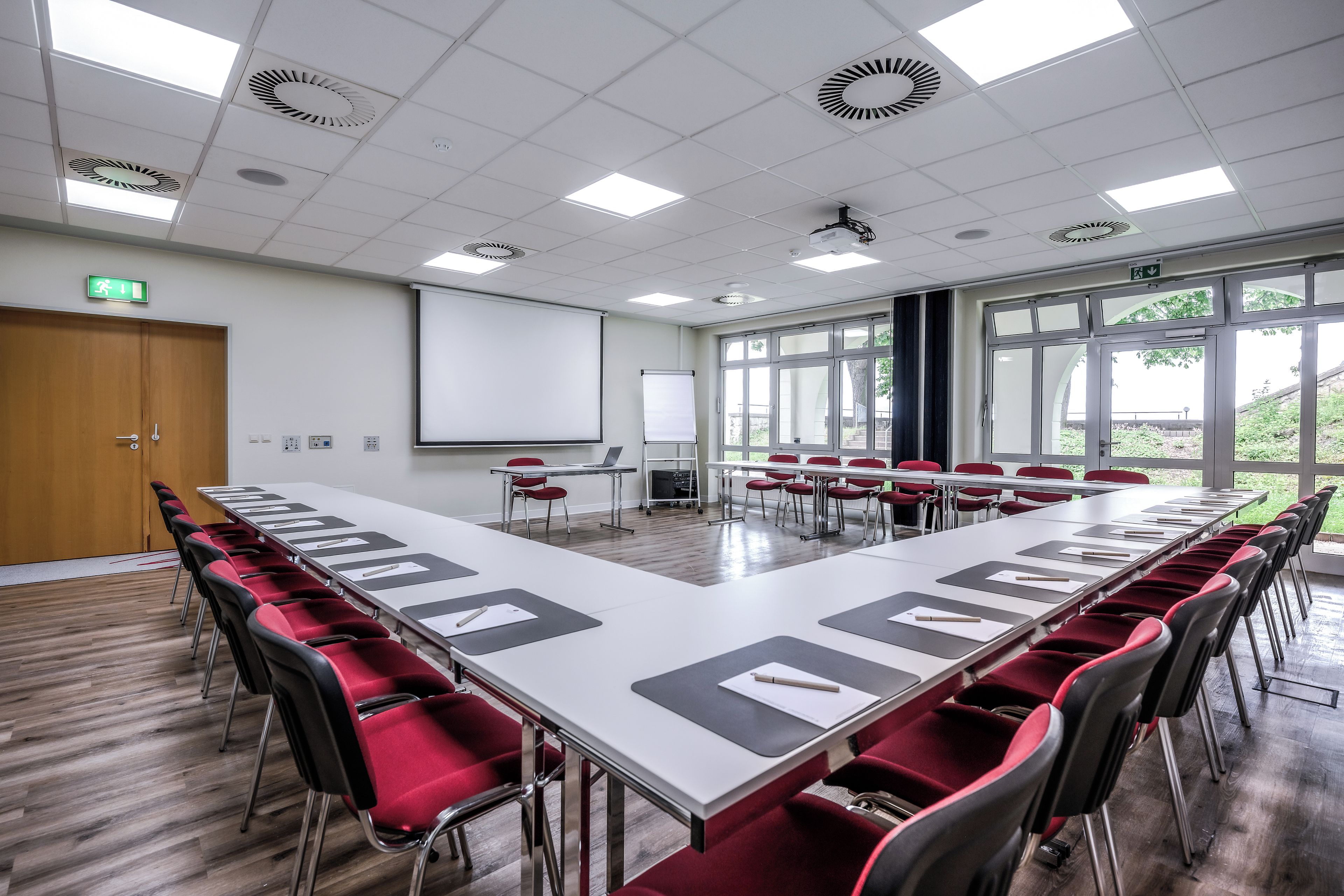 Un salón de conferencias con una larga mesa, sillas rojas, una pantalla para proyector y una pizarra.