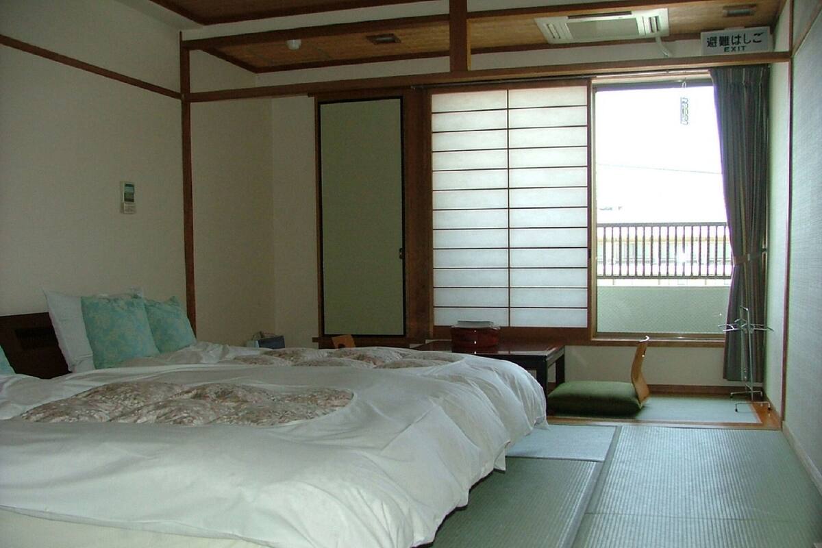 Une pièce de style japonais traditionnel, avec un sol recouvert de tatamis, un grand lit avec une literie blanche, une porte coulissante de type shoji et une vue sur un balcon.
