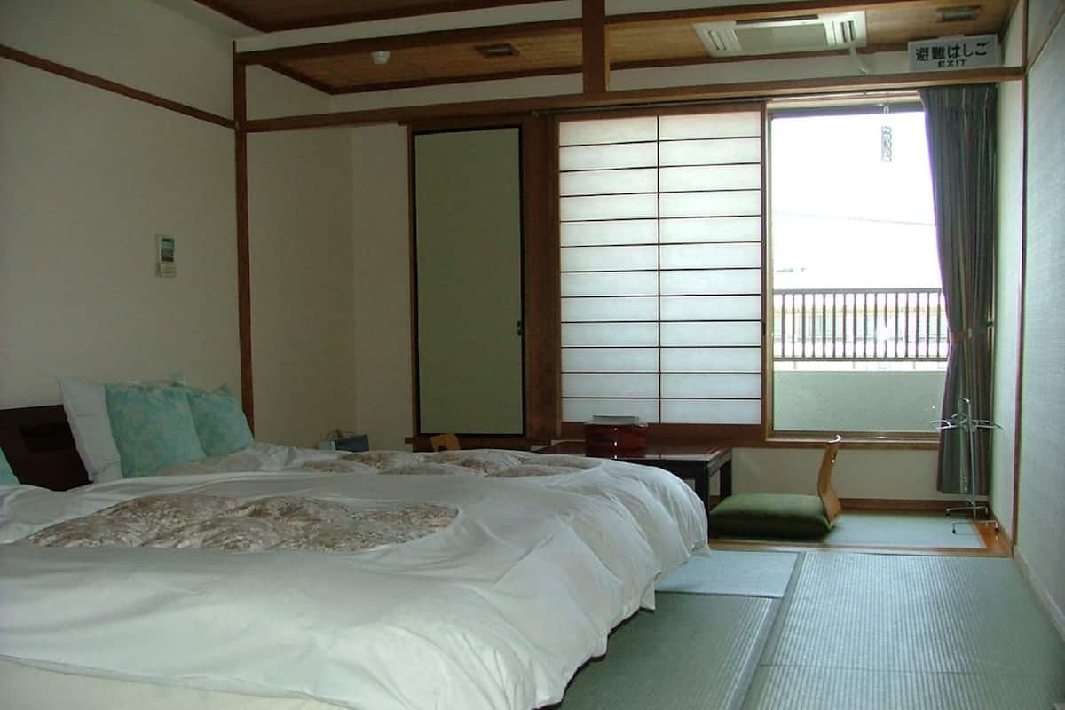 Una habitación japonesa tradicional con suelo de tatami, una cama grande con ropa de cama blanca, una puerta corrediza de shoji y vistas a un balcón.
