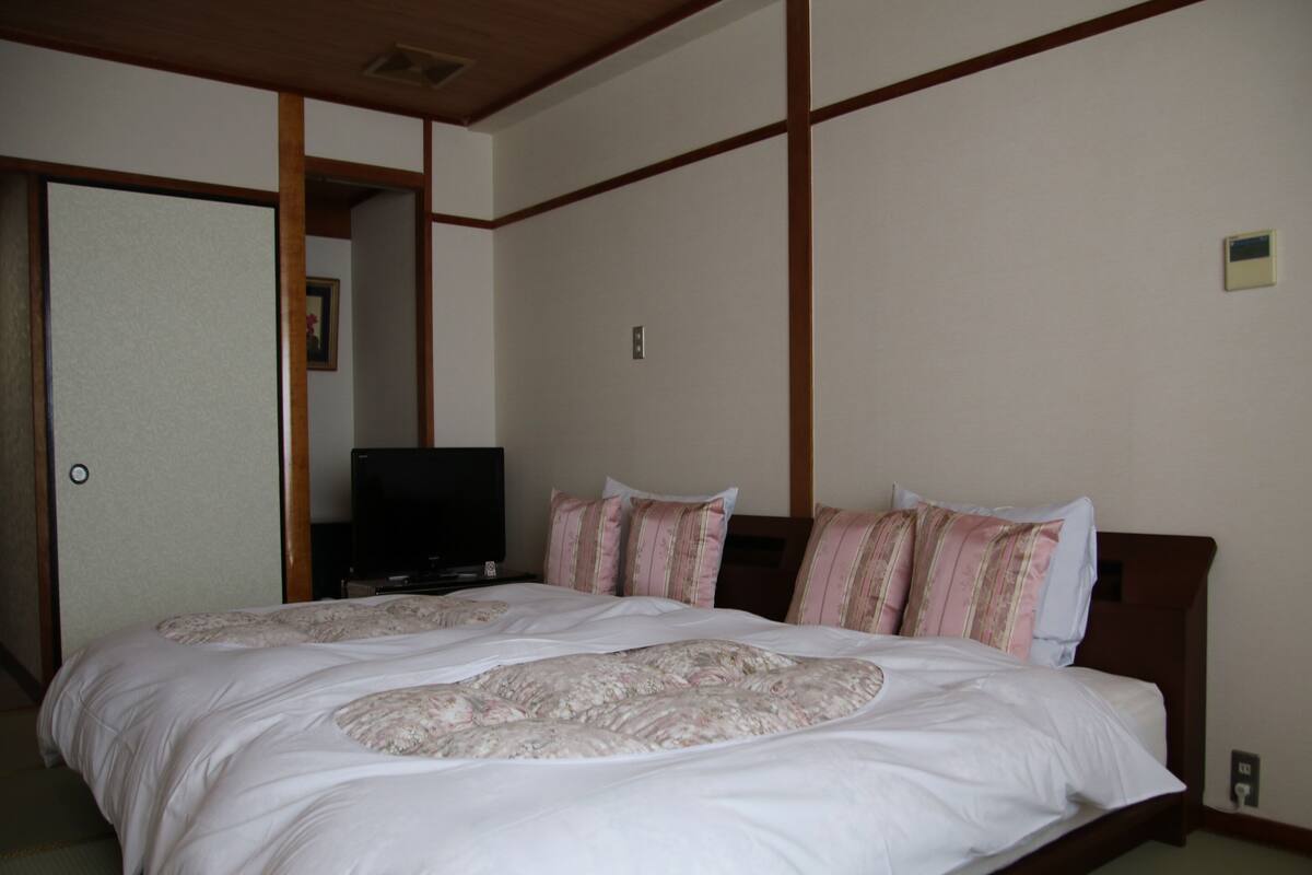 Un lit bien fait, recouvert d’une couvre-lit à motifs floraux, un téléviseur posé sur un meuble et une tête de lit en bois.